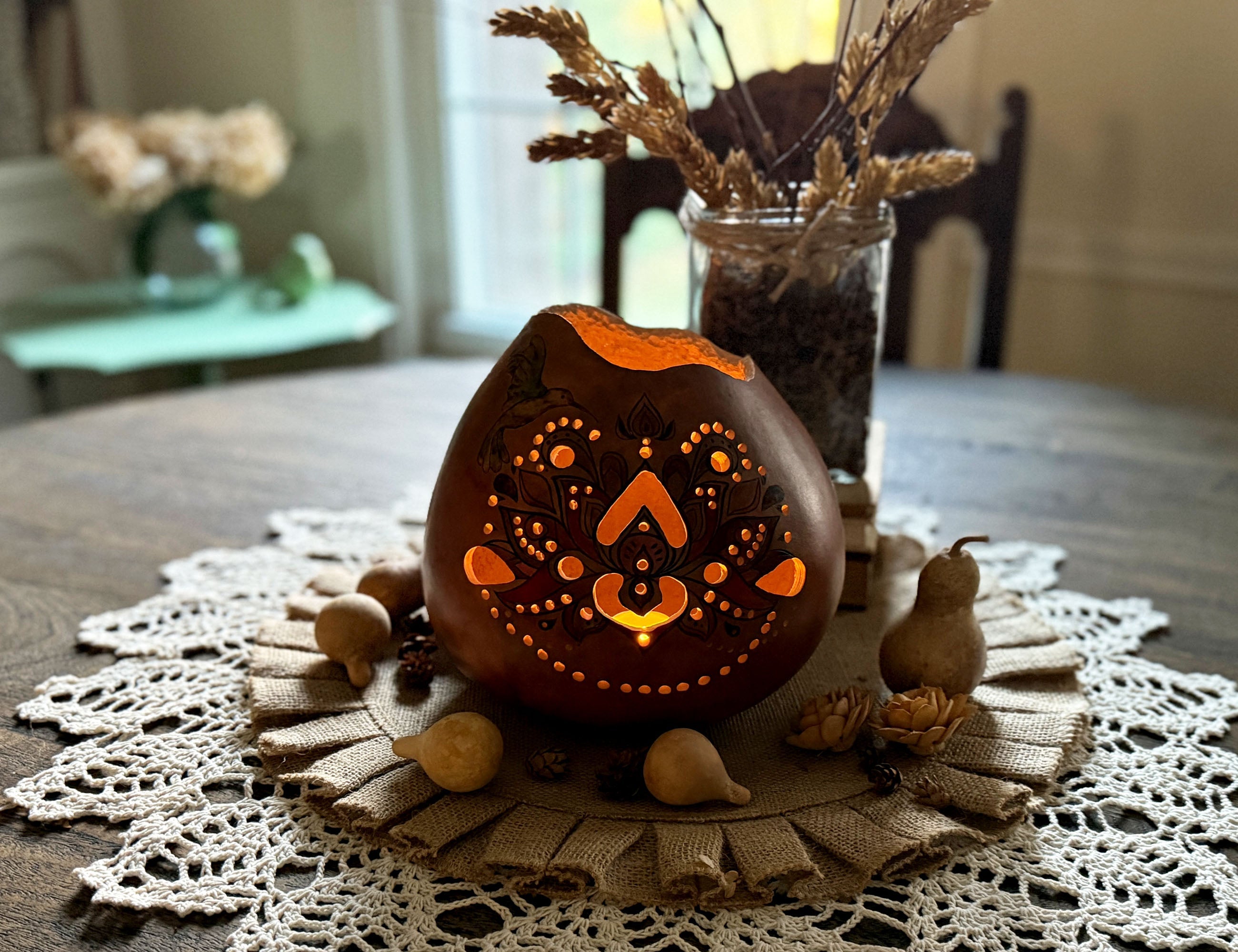 A hand carved gourd luminary sits on a round wood dinning room table. A candle lights the mandala design cut out of the gourd that is sitting on a crochet doily with a vase of hops behind it.