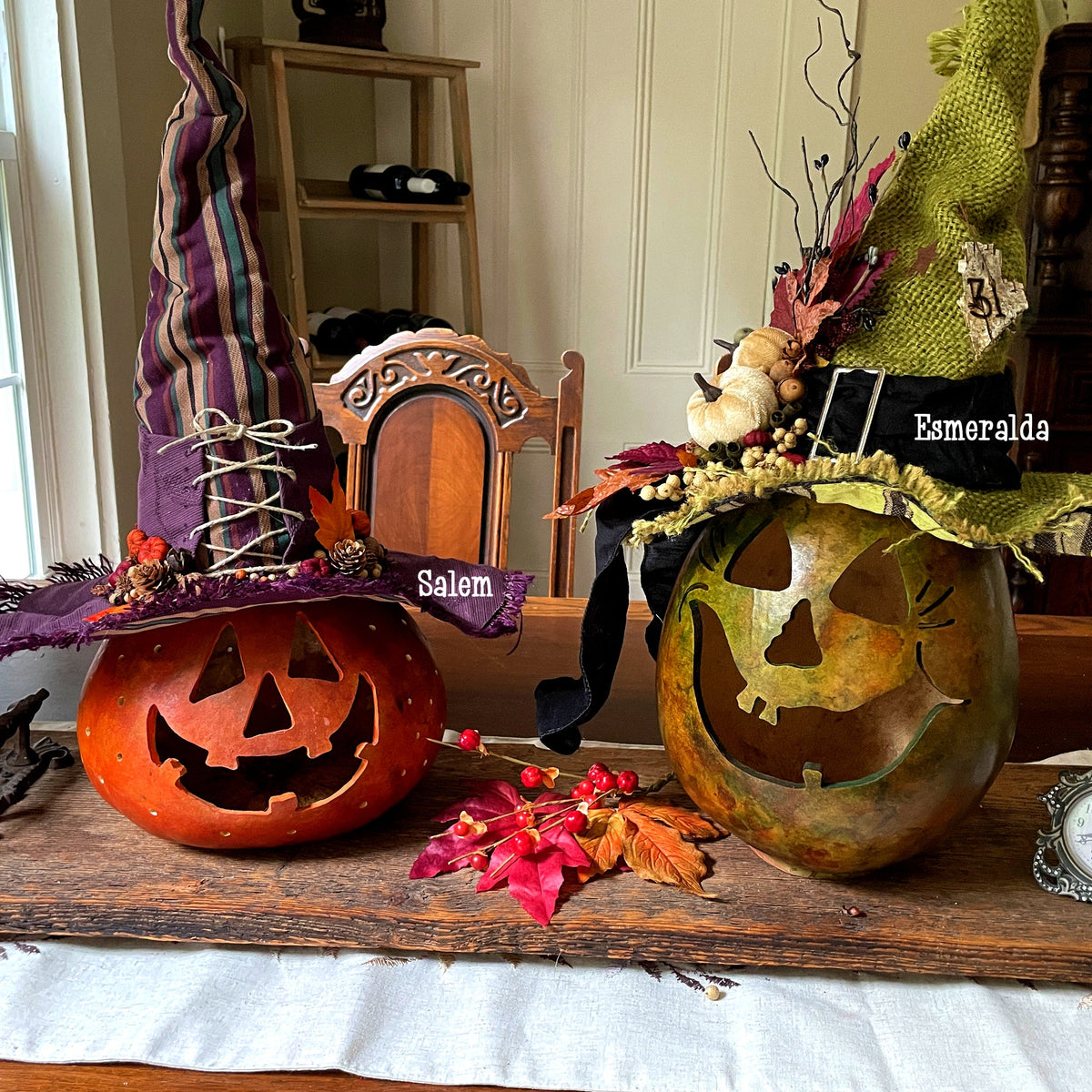 Two extra large pumpkin gourds sit on a dinning room table scape. They are hand carved, one stained orange the other stained green. Each pumpkin has it's own custom made witch hat.
