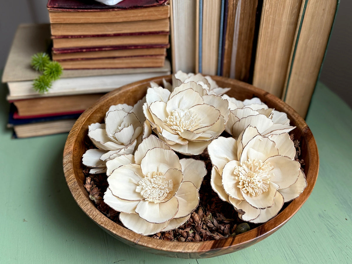 A round wooden bowl with stacked vintage books behind it. The bowl holds and arrangement of cedar cone filler and several wood flowers blossoms in a neutral beige. 