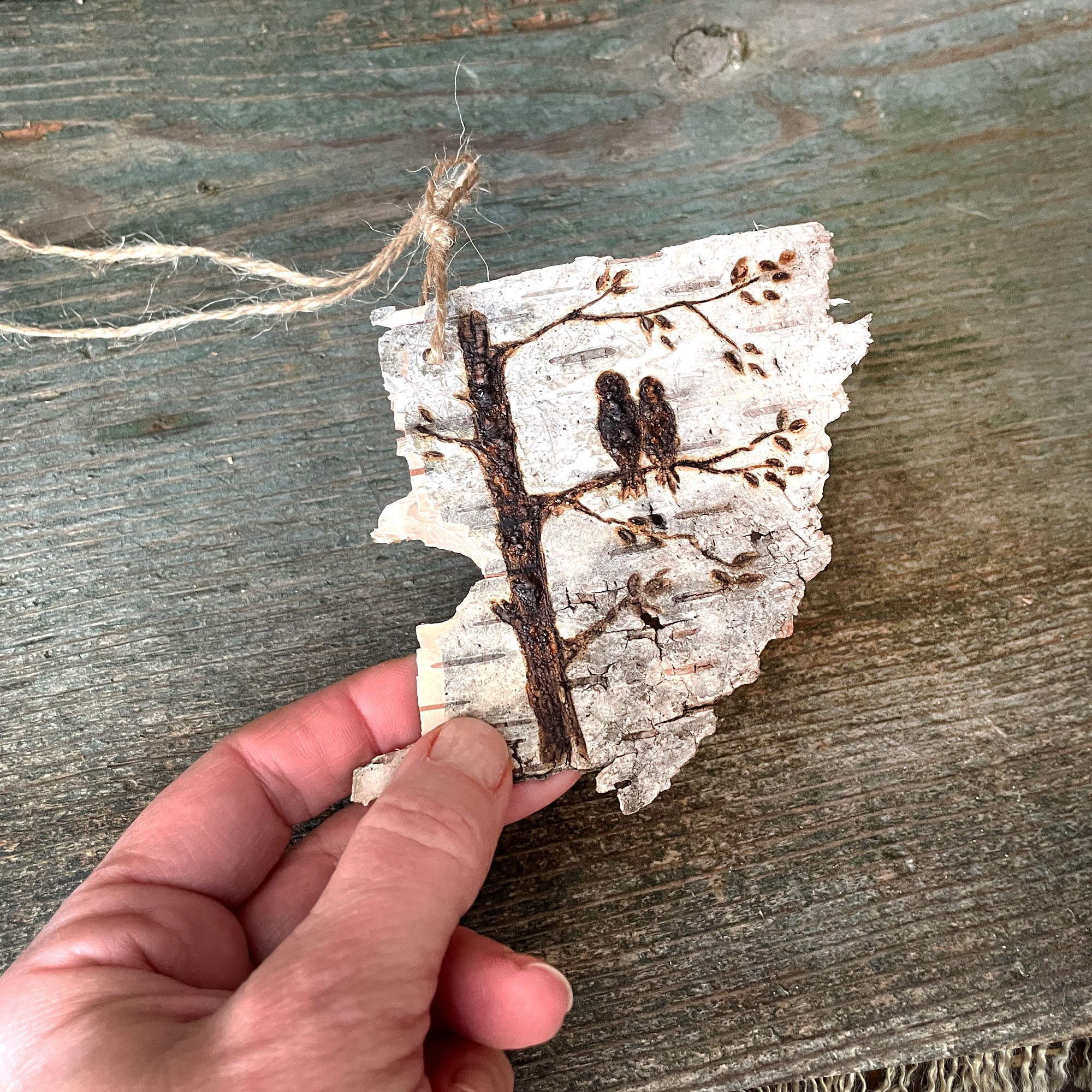 Me holding a real piece of birch bark. It's a three inch torn piece with a jut cord tied to the top. I have hand drawn two birds sitting on a tree branch using pyrography or wood burning.