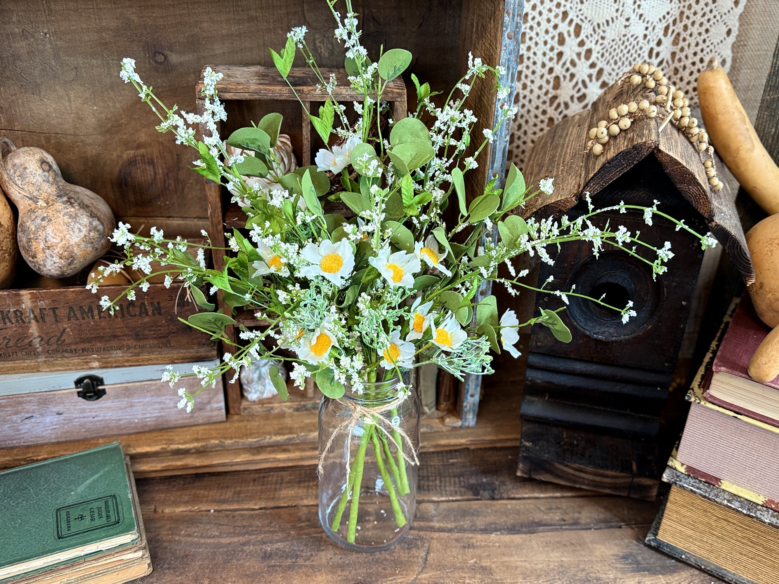 A rustic centerpiece featuring a faux flower arrangement of a white daisies and green eucalyptus. The flowers are in a mason jar. The jar sits on a  old wood table. A brown bird house sites in the background.