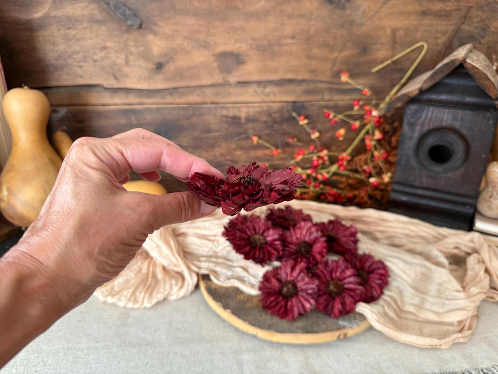 Hand holding dried red flowers over a woven basket with more flowers on a wooden surface.