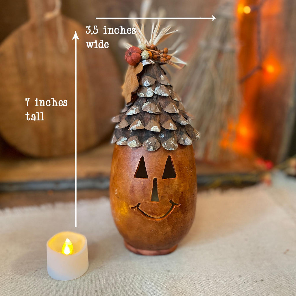 Decorative bottle with a pumpkin face and pine cone top, labeled for width and height, next to a candle.