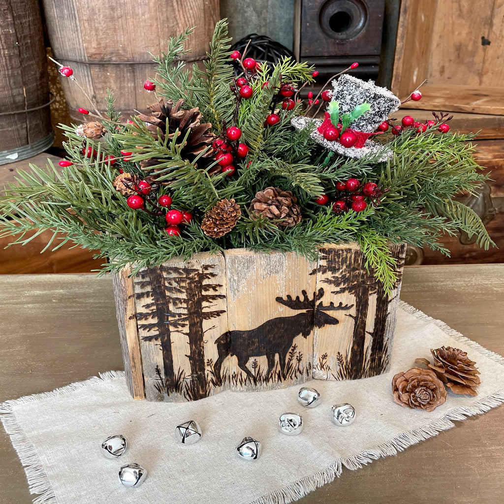 Decorative Christmas arrangement with greenery, berries, and pinecones in a wooden box with moose design on a wooden surface.