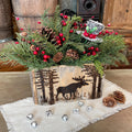 Decorative Christmas arrangement with greenery, berries, and pinecones in a wooden box with moose design on a wooden surface.