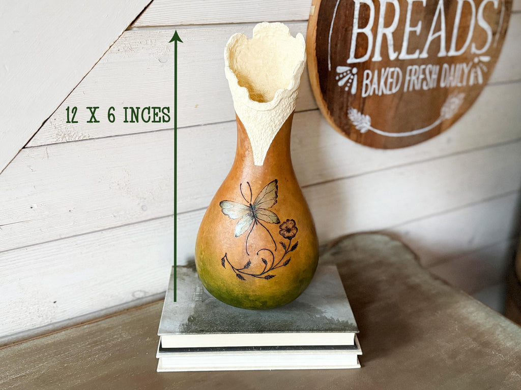Decorative gourd with butterfly design on a wooden surface, with a 'Breads Baked Fresh Daily' sign in the background.