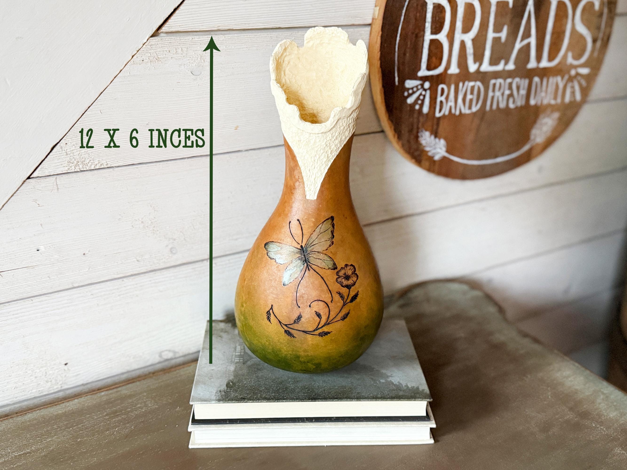 Decorative gourd with butterfly design on a wooden surface, with a 'Breads Baked Fresh Daily' sign in the background.