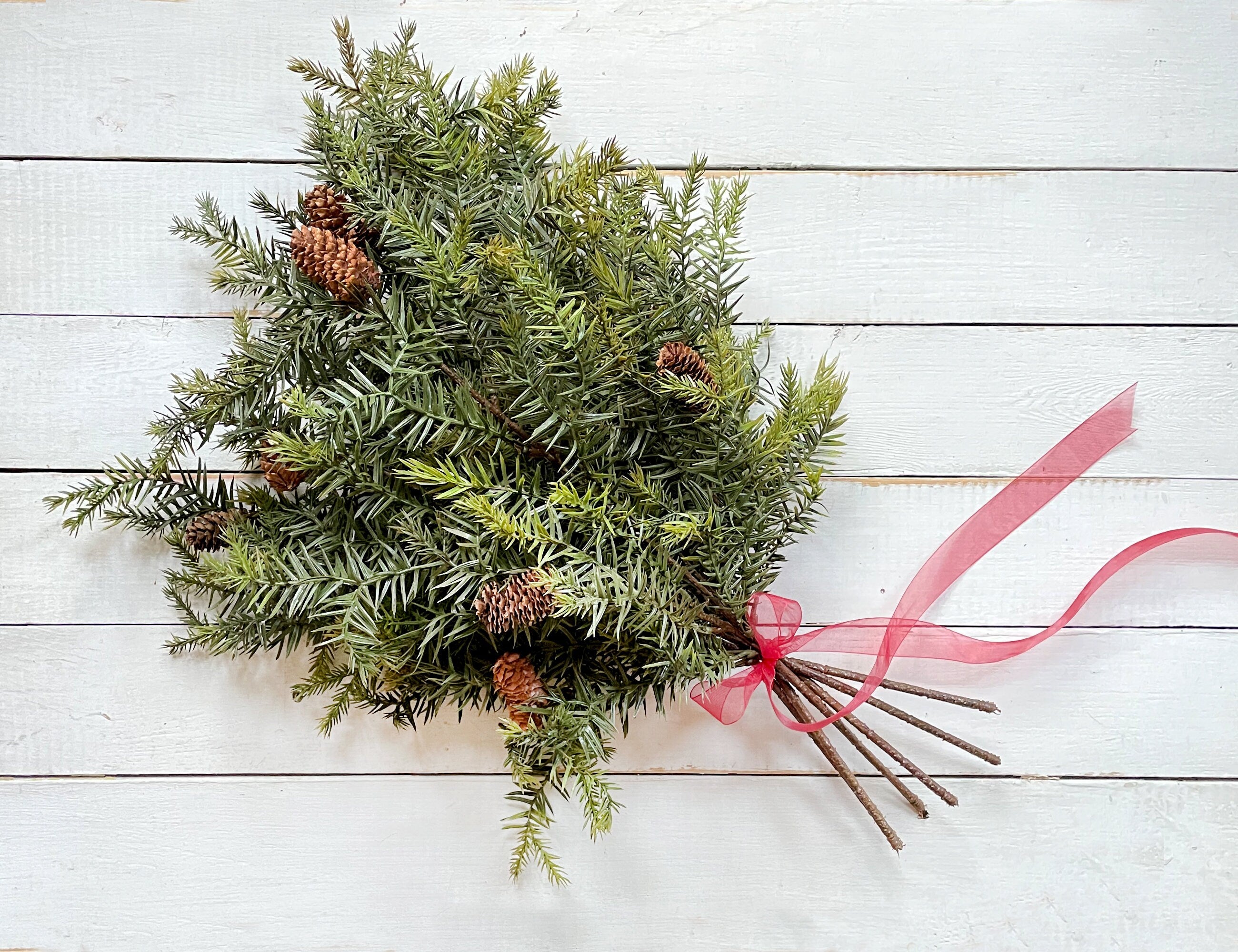 Greenery bouquet with pinecones and a pink ribbon on a white wooden background