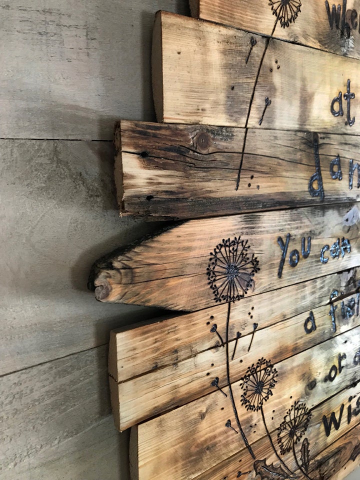 Wooden wall art with engraved text and dandelion designs on a concrete wall.