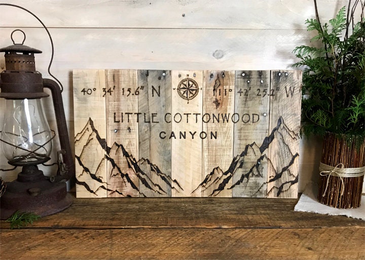 Wooden sign with mountain design and text 'Little Cottonwood Canyon' on a wooden surface with lantern and plant.
