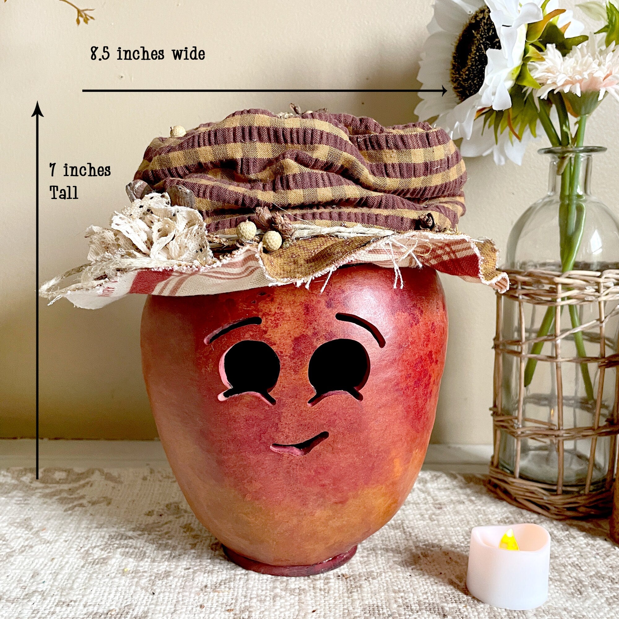 Decorative apple-shaped container with face details on a surface, with measurements and a candle in the background.