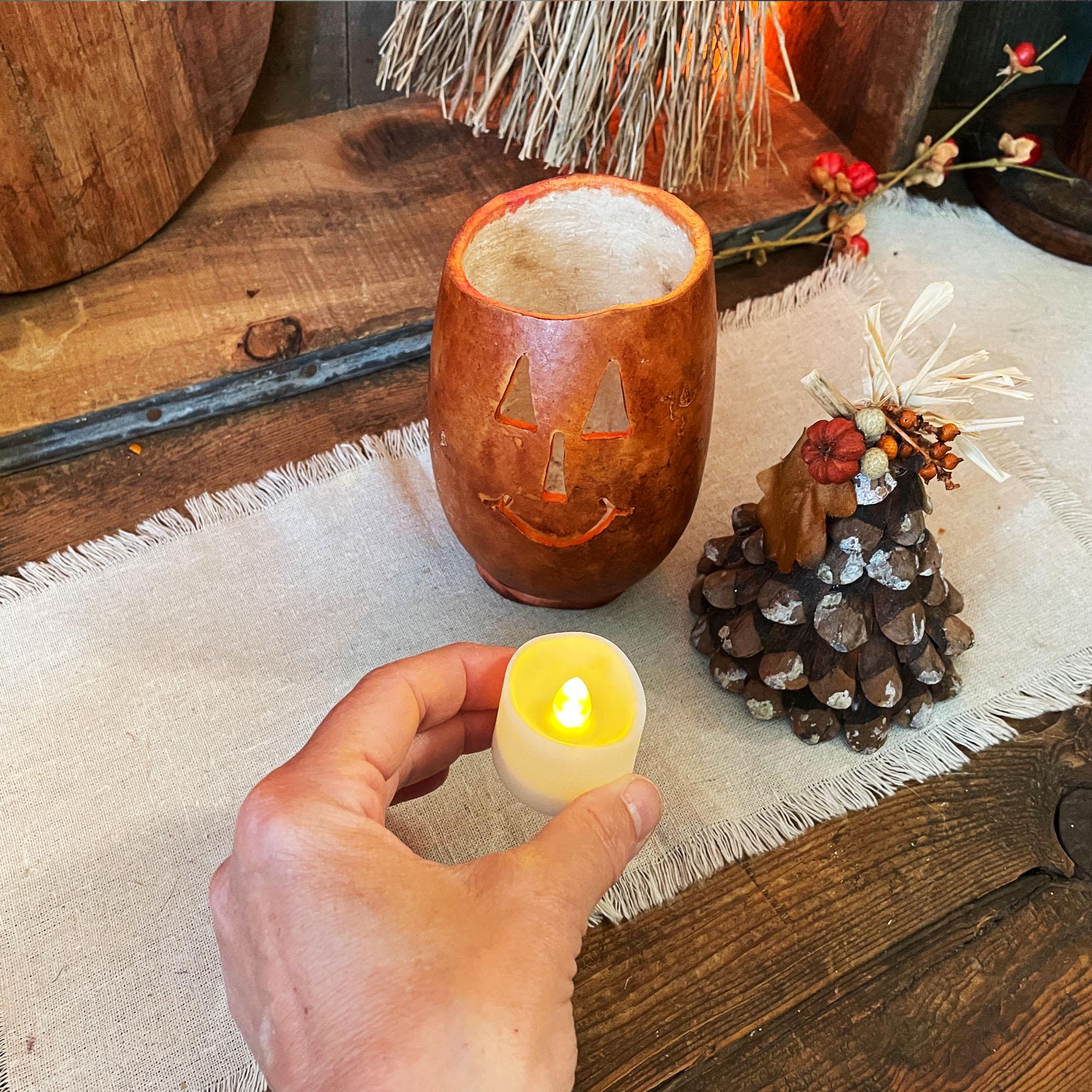 Hand holding a small lit candle next to a carved pumpkin lantern and decorative pine cone on a wooden surface.