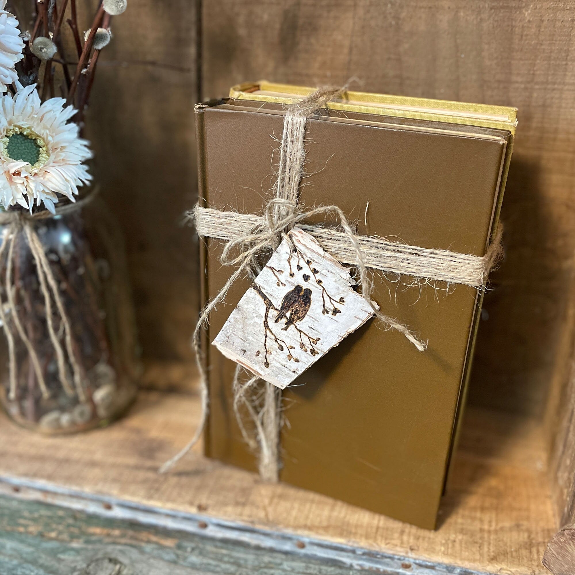 Gold-bound book tied with twine and a decorative tag on a wooden surface with flowers in the background
