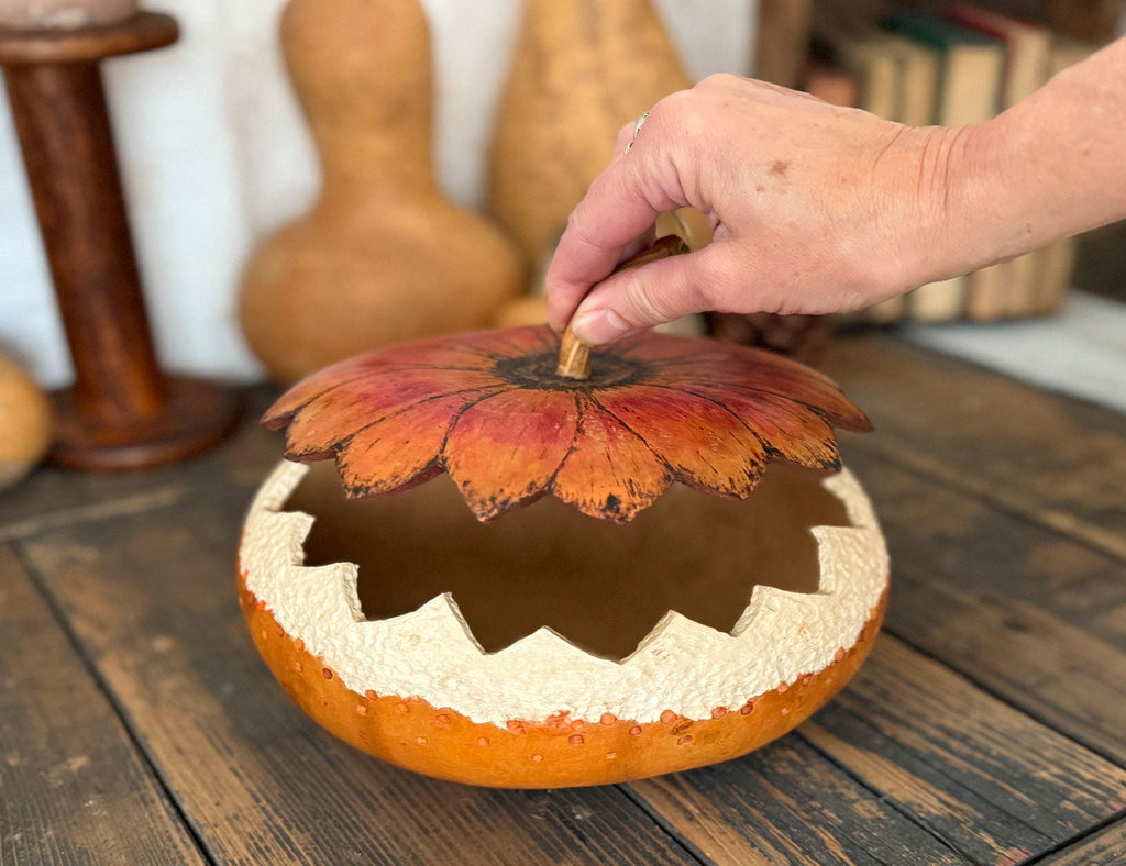 Decorative gourd with floral design with lid being adjusted by a hand on a wooden surface.