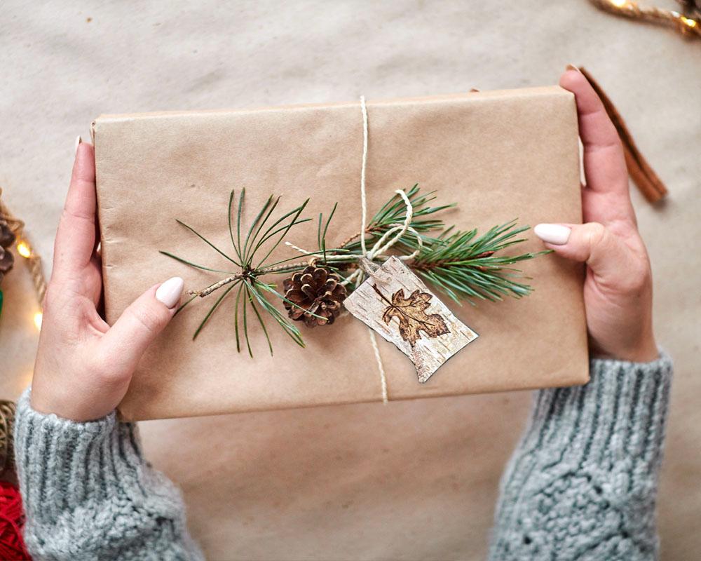 Woodland Gift wrapped in brown kraft paper with a pine branch and decorative tag, held by hands wearing gray sleeves.
