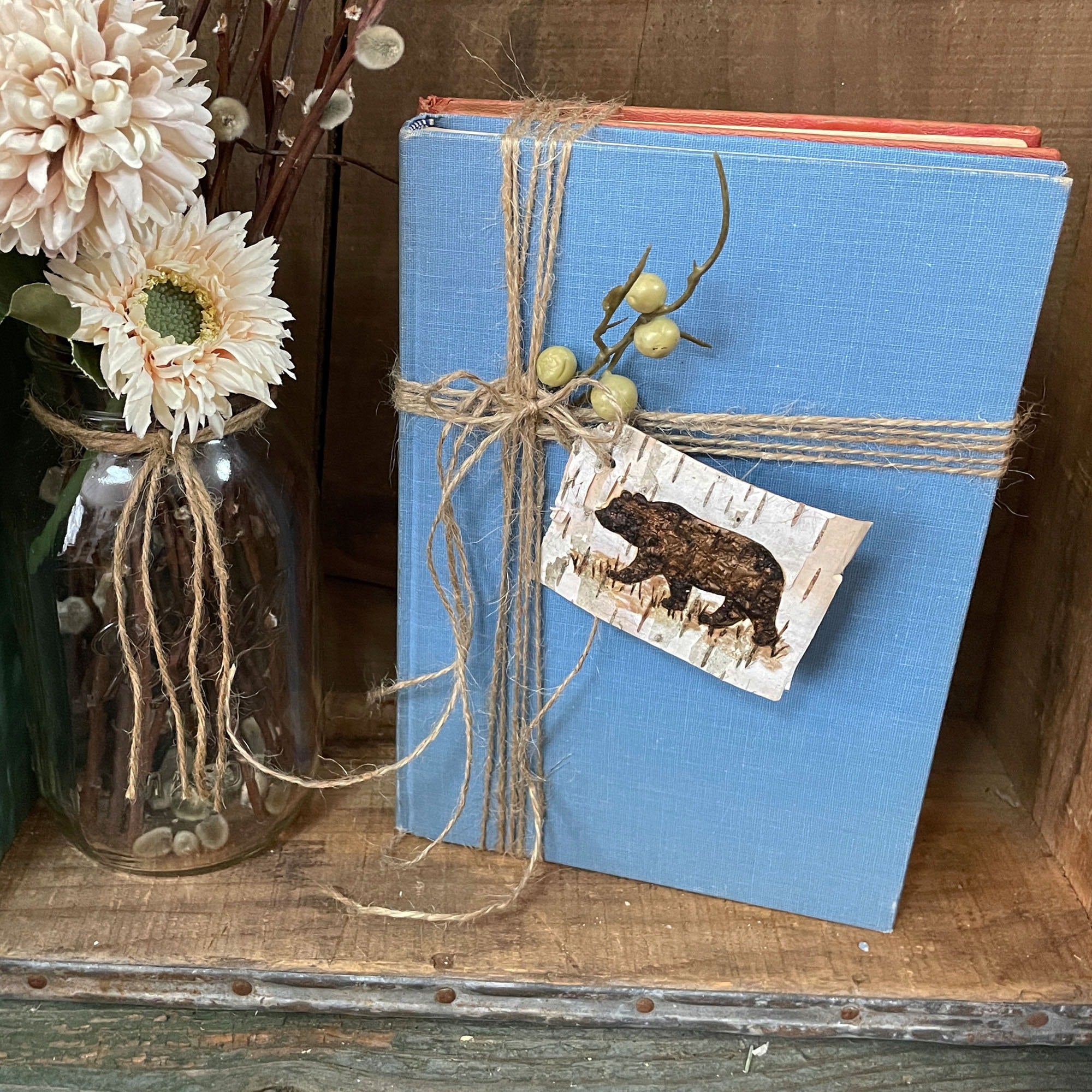 Blue book tied with twine and a bear accent tag on a wooden surface with flowers.