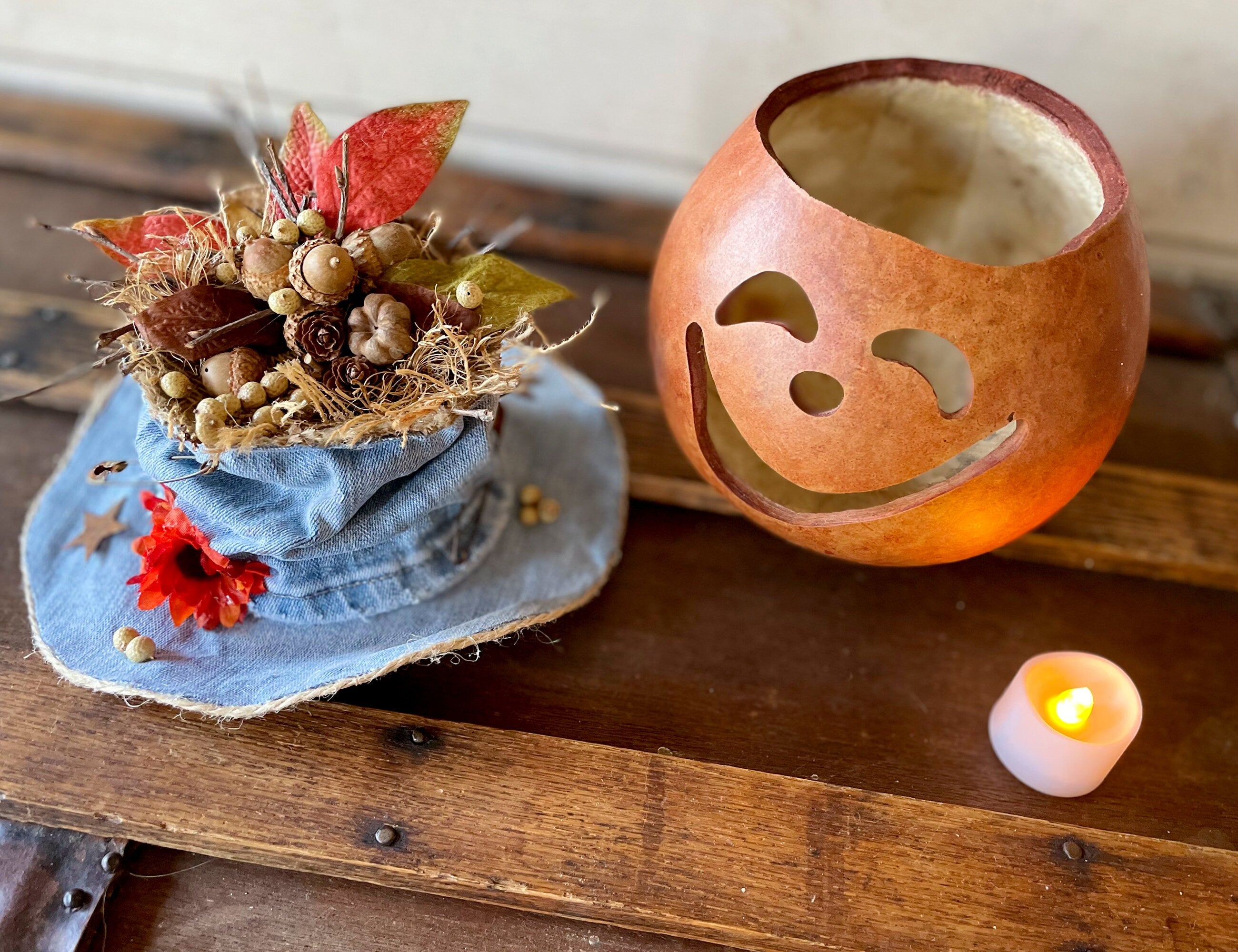 Decorative pumpkin with face carving, small hat with flowers, and candle on a wooden surface