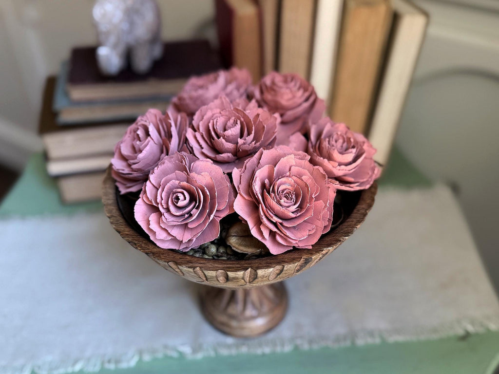 Decorative arrangement of pink wood flowers in a wooden bowl on a table with books in the background.