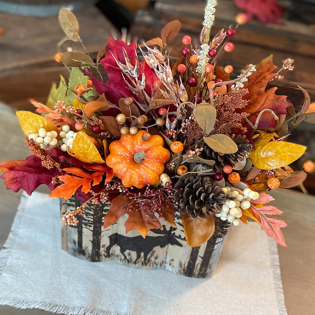 Autumn-themed floral arrangement with pumpkins and leaves in a wooden box on a white cloth.