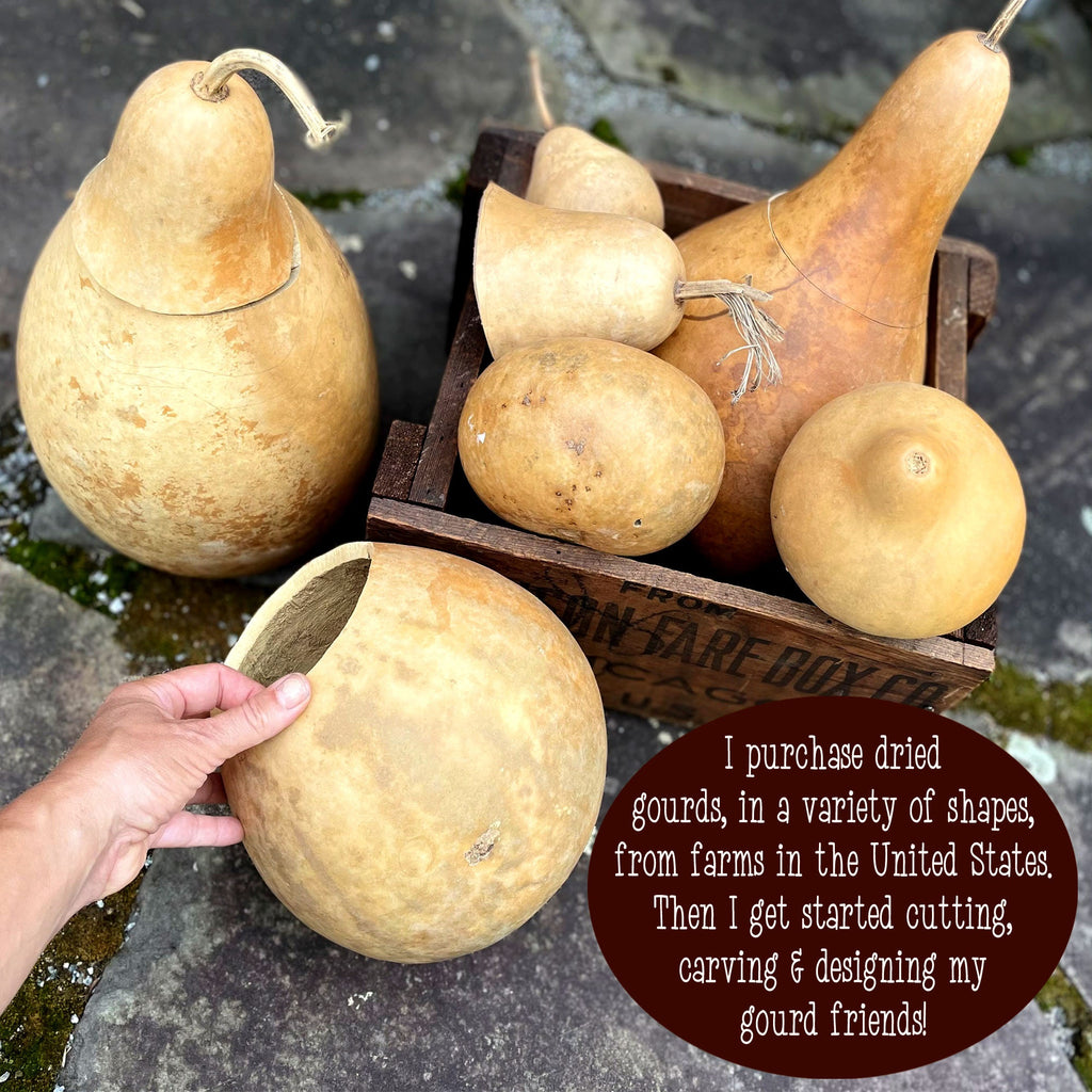 Hand holding a dried gourd with other gourds in a wooden crate on a stone surface.