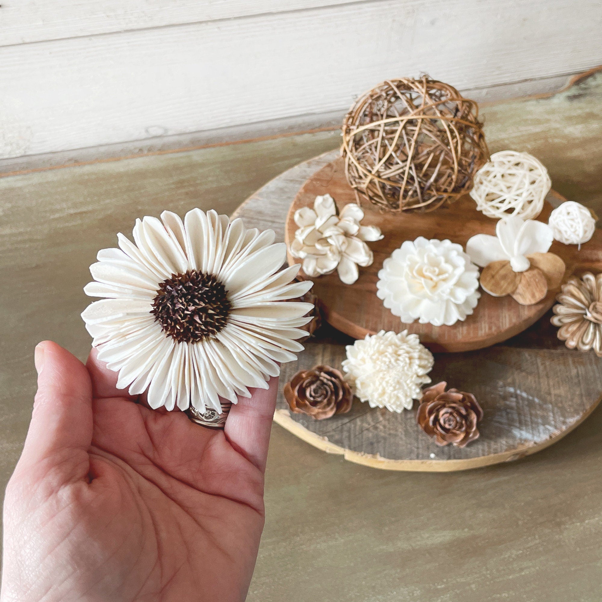 Hand holding a white flower-shaped object with a wooden surface and decorative items in the background.