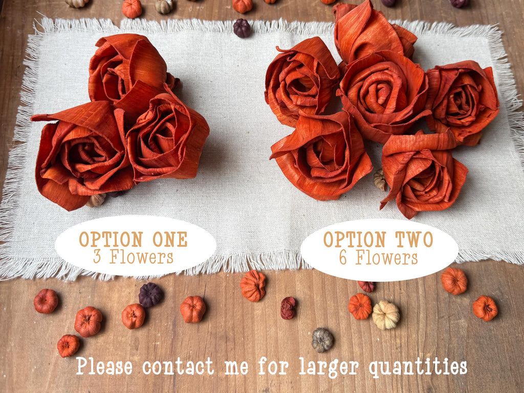 Two options of orange fabric flowers on a white cloth with additional flowers on a wooden surface.