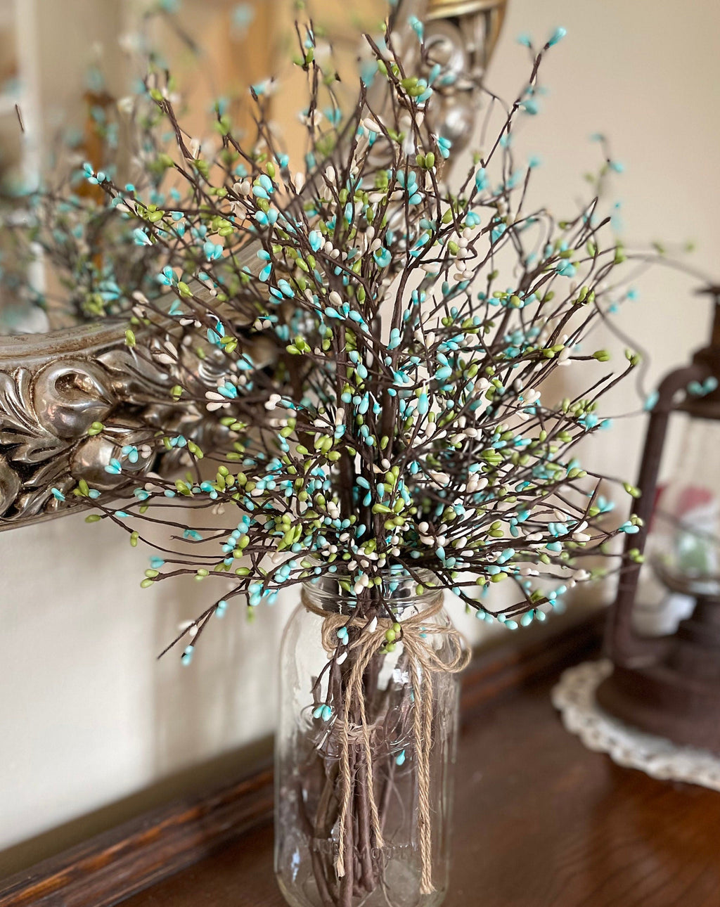 Decorative arrangement of branches with turquoise beads in a glass jar, placed on a surface with a mirror and other items in the background.