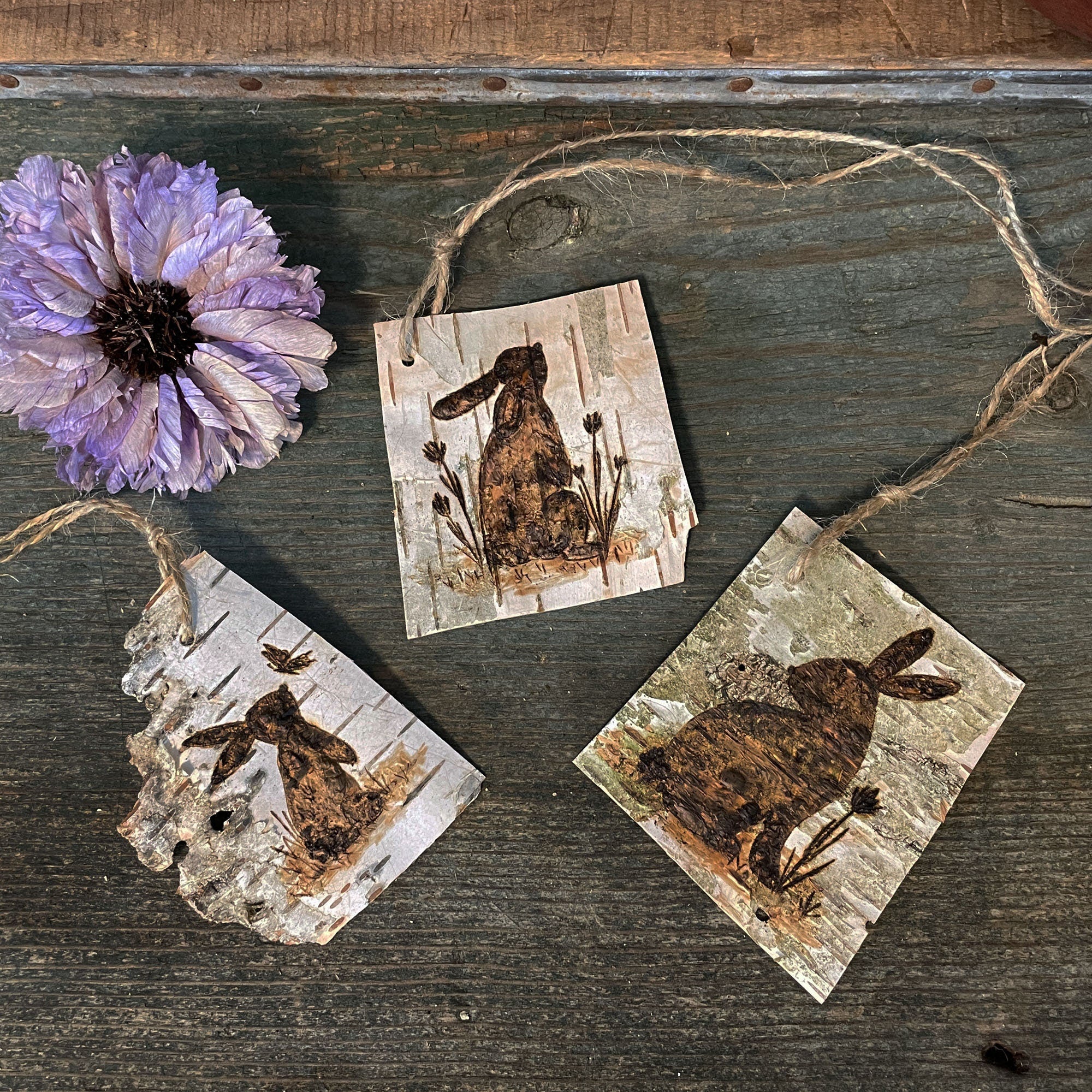 Three square white birch bark gift toppers with rabbit designs on a wooden surface with a purple flower.