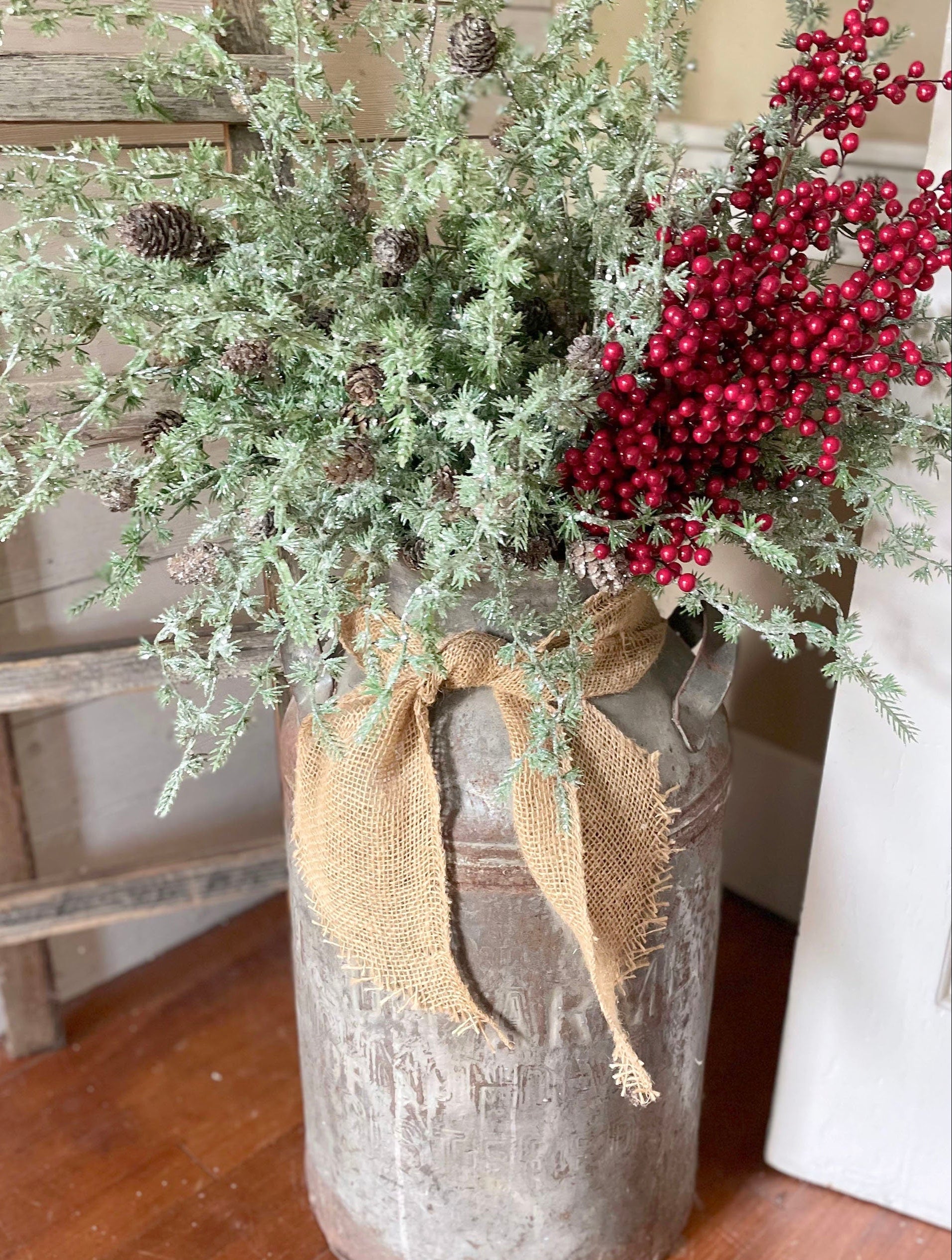 Decorative arrangement with a milk can, greenery, and red berries on a wooden floor.