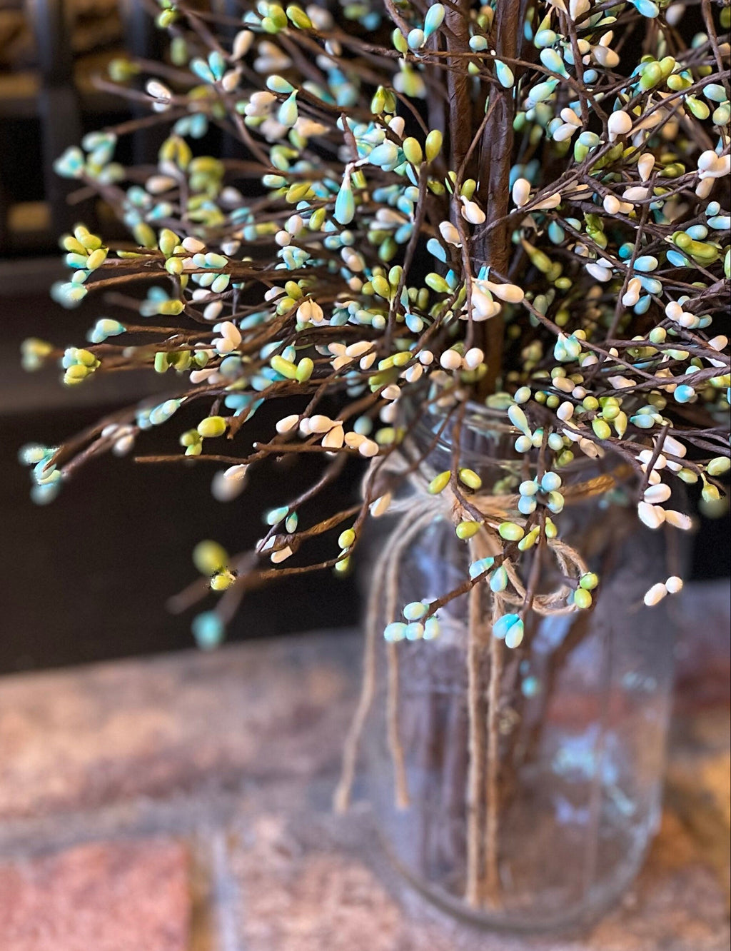 Decorative arrangement of branches with small green and blue berries in a clear glass jar on a brick surface.