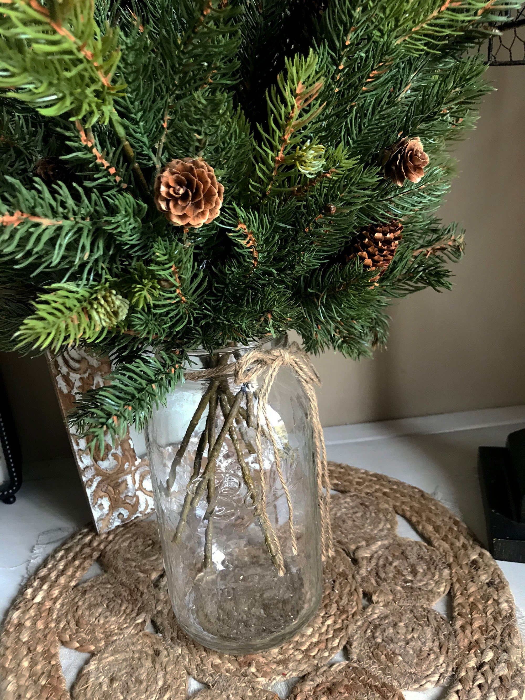 Decorative arrangement of greenery and pinecones in a glass vase on a textured surface.