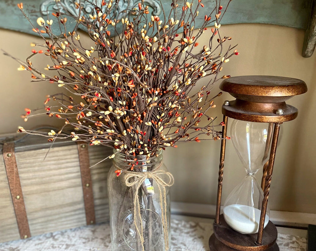 Decorative arrangement with a jar of branches and berries next to an hourglass on a textured surface.