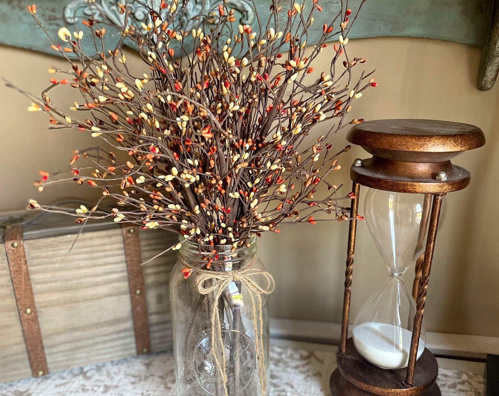 Decorative arrangement with a jar of branches and berries next to an hourglass on a textured surface.
