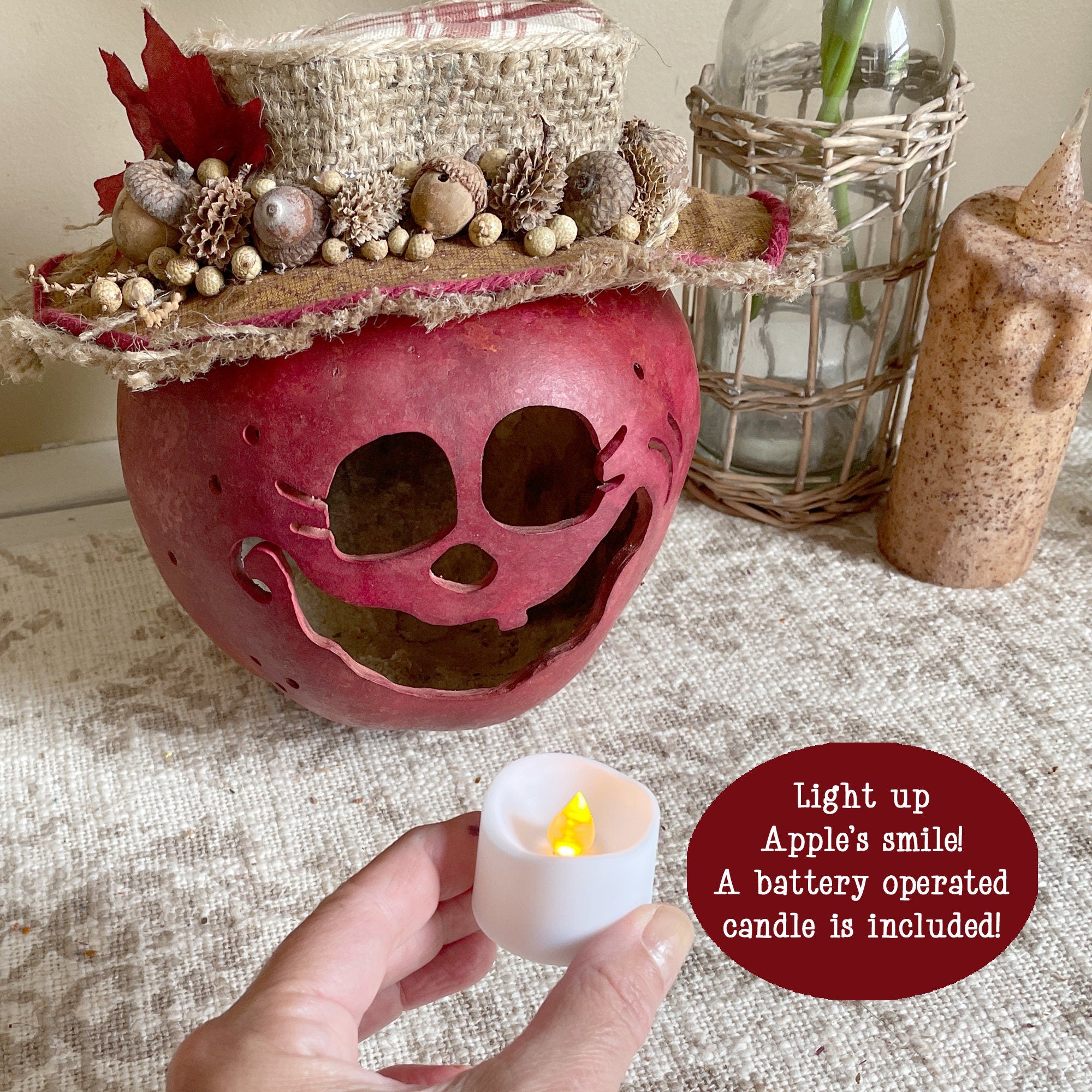 Decorative apple with a carved face, topped with autumnal elements, held by a hand with a battery-operated candle.
