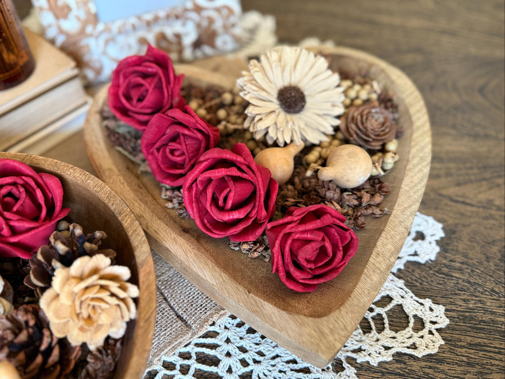 Heart-shaped wooden box filled with red roses, pinecones, and a white flower on a lace doily.