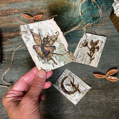 Hand holding a woodland birch bark tag with engraved fairy design on a wooden surface.