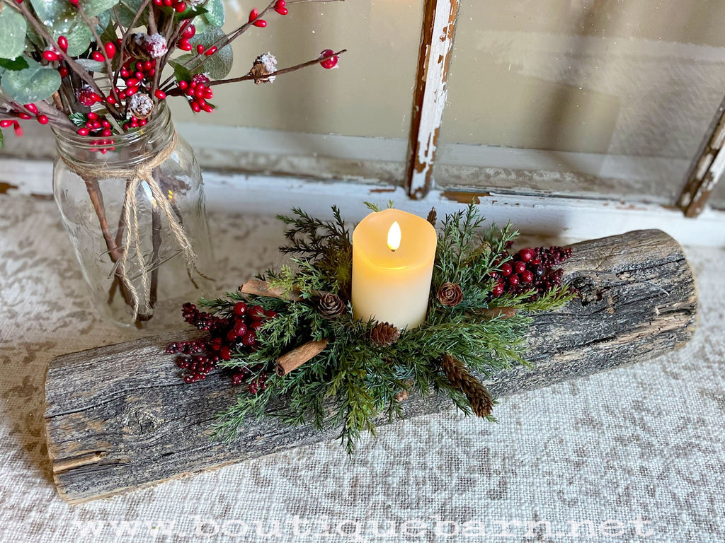 Decorative setup with a candle on a log, surrounded by greenery and berries, against a neutral background.