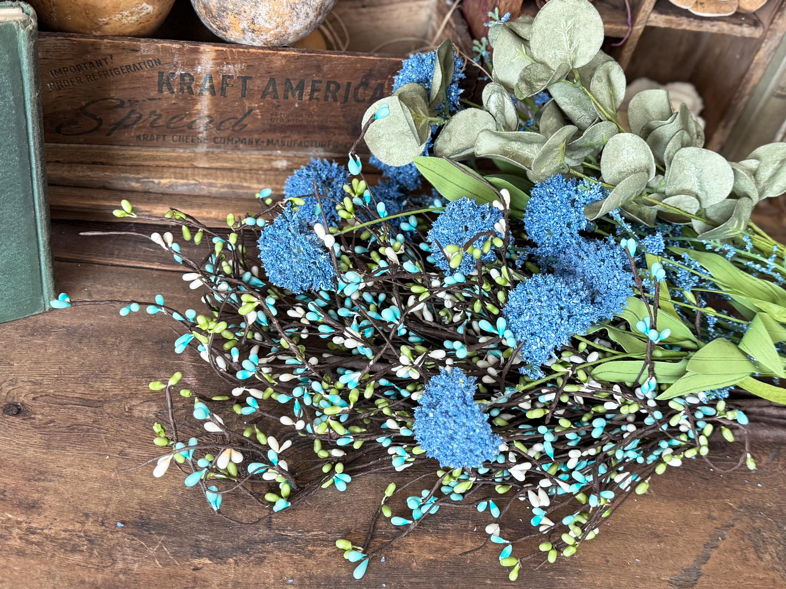 Decorative arrangement of blue flowers and greenery on a wooden surface with a wooden box labeled 'Kraft America' in the background.