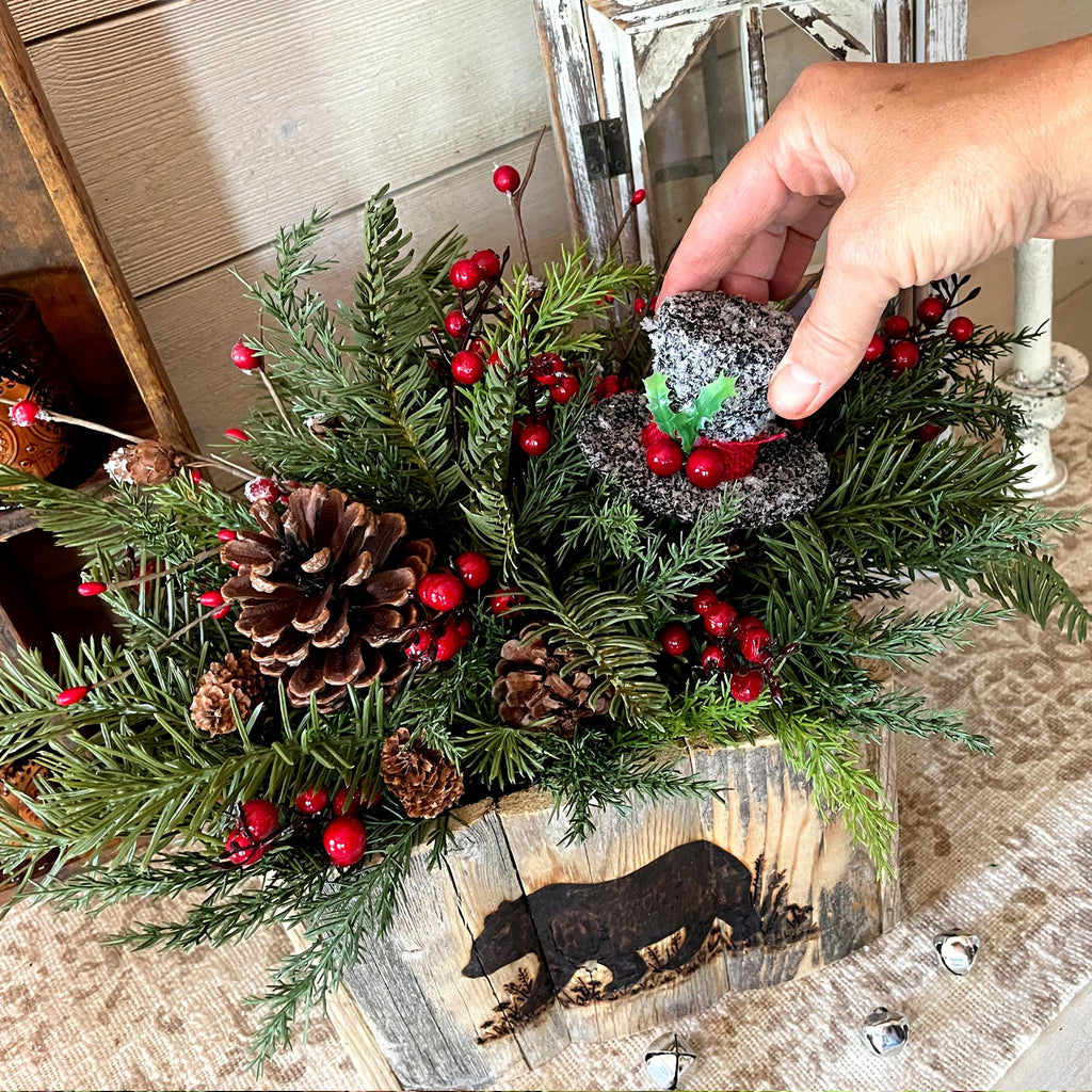 Hand arranging a festive wreath with pine cones, berries, and a small figurine on a wooden surface.