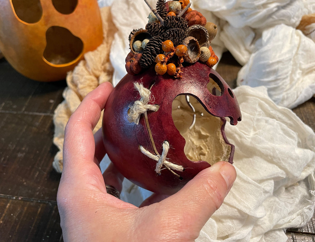 Decorative pumpkin with a carved face held by a hand, surrounded by white fabric and a pumpkin in the background.