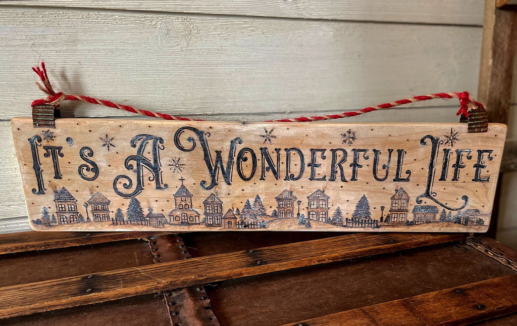 Wooden sign with 'It's a Wonderful Life' text and house illustrations on a rustic wooden surface.