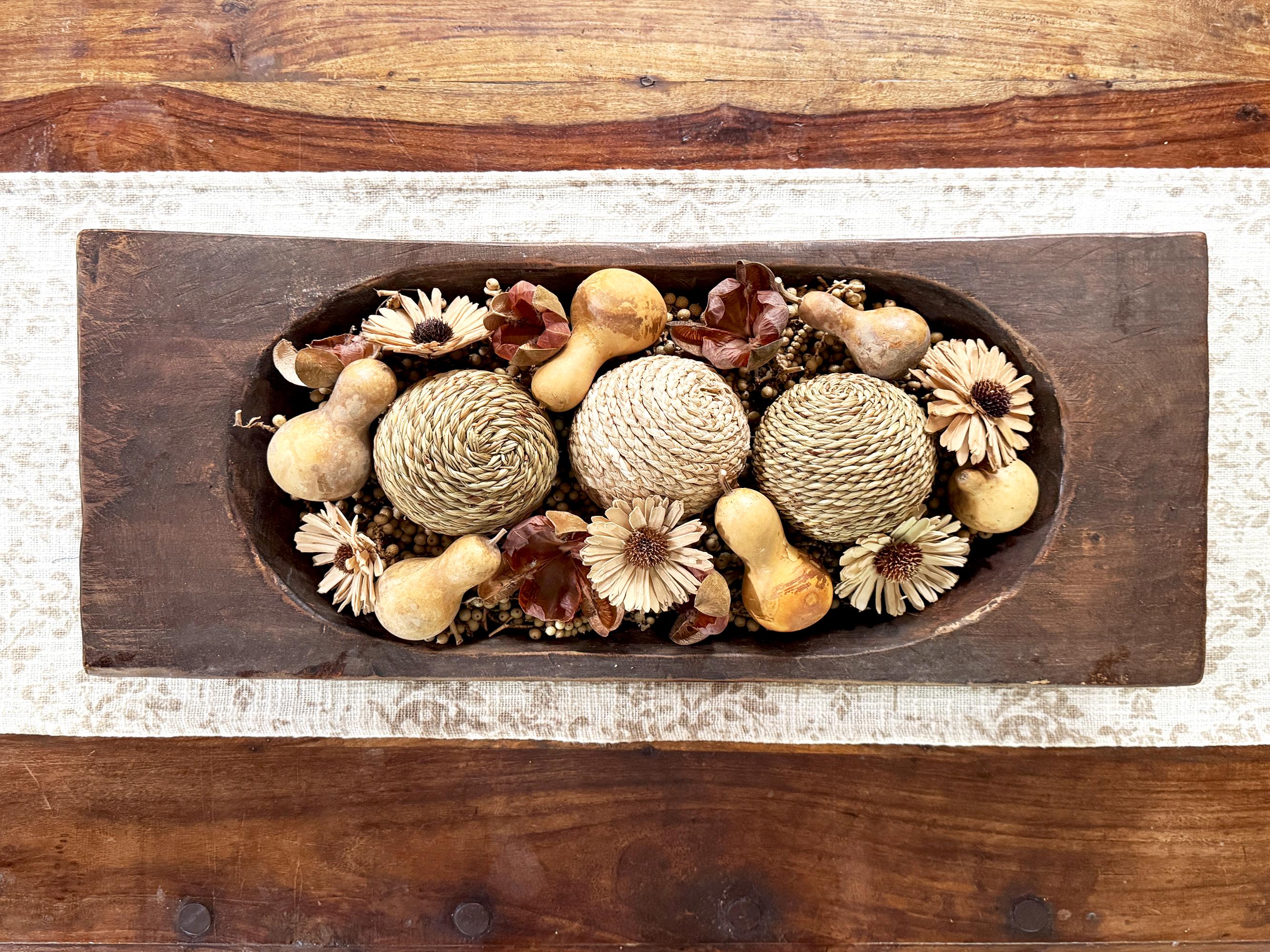 Decorative arrangement of gourds and flowers in a wooden dough bowl on a coffee table runner.