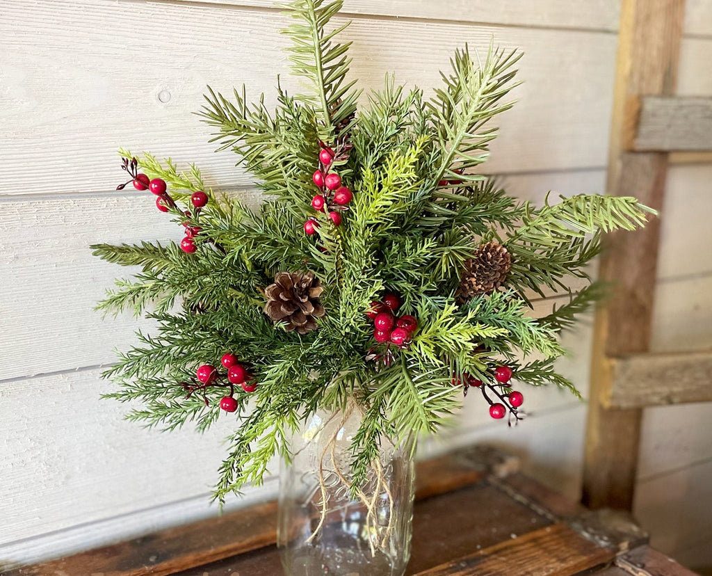 Decorative arrangement of greenery with red berries and pinecones in a clear vase against a wooden background.
