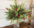 Decorative arrangement of greenery with red berries and pinecones in a clear vase against a wooden background.