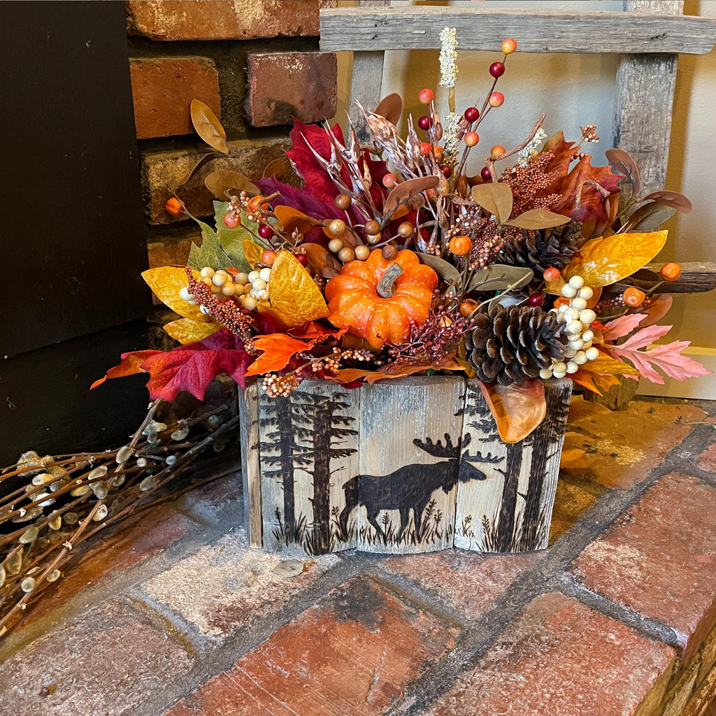 Decorative fall arrangement with pumpkins and leaves in a wooden box with a moose design on a brick surface.