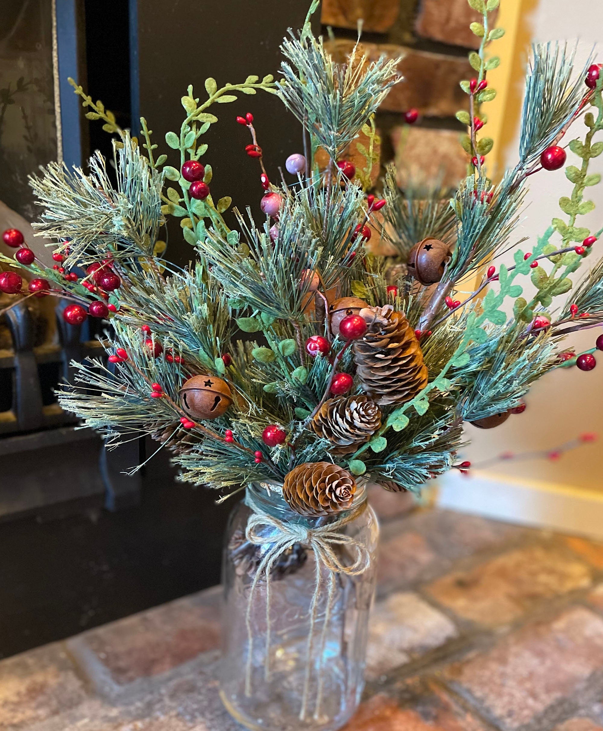 Decorative arrangement of pine branches, red berries, and pinecones in a glass jar on a stone surface.