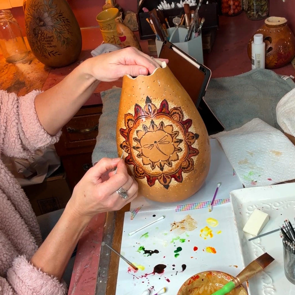 Person painting a decorative design on a gourd with various art supplies around.