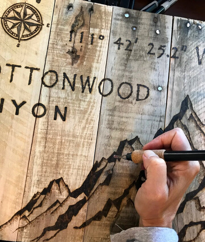 Hand holding a tool against a wooden sign with 'Cottonwood Canyon' and mountain designs.