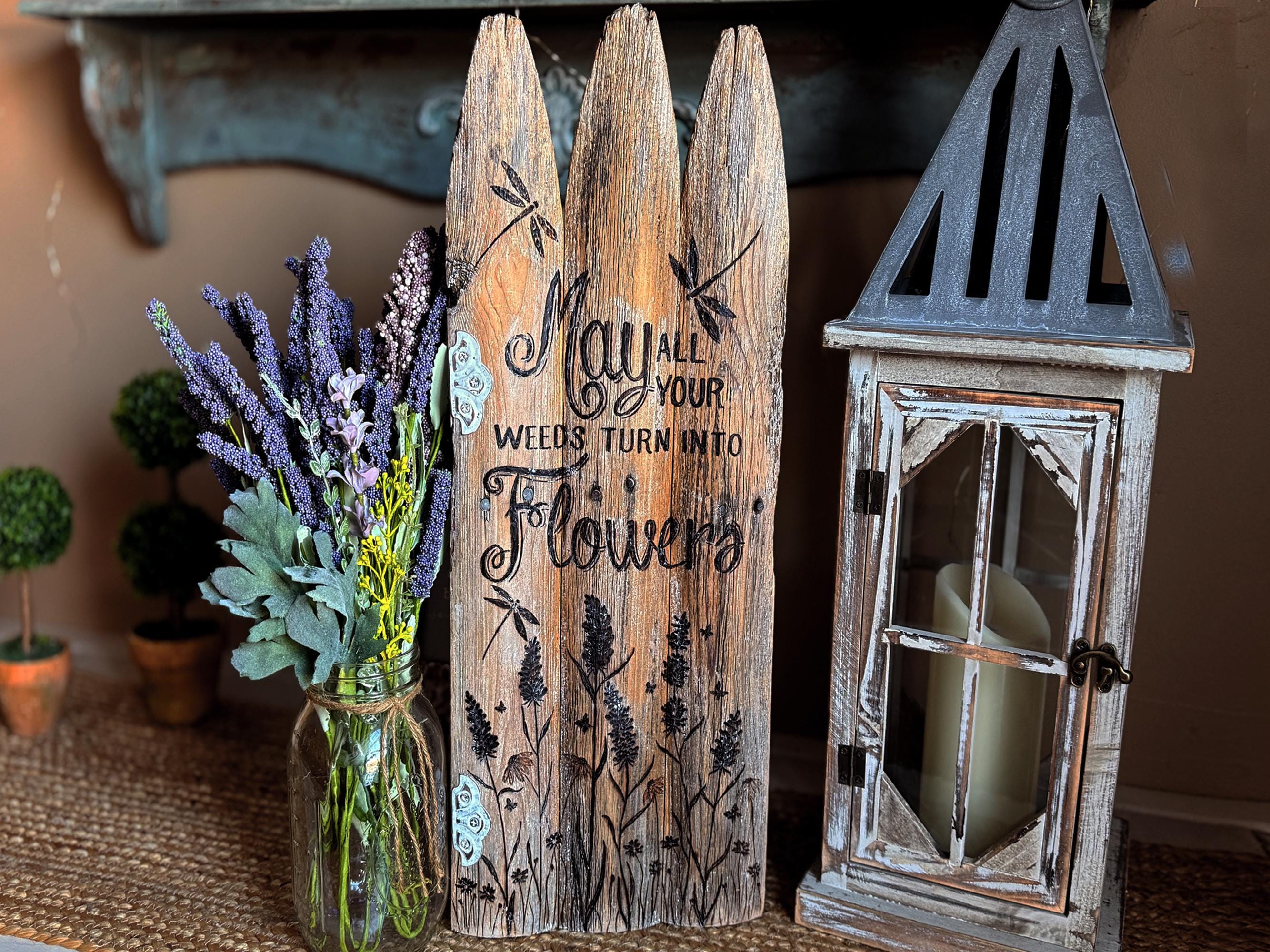 Decorative wooden sign with floral design and lantern next to a vase with flowers.