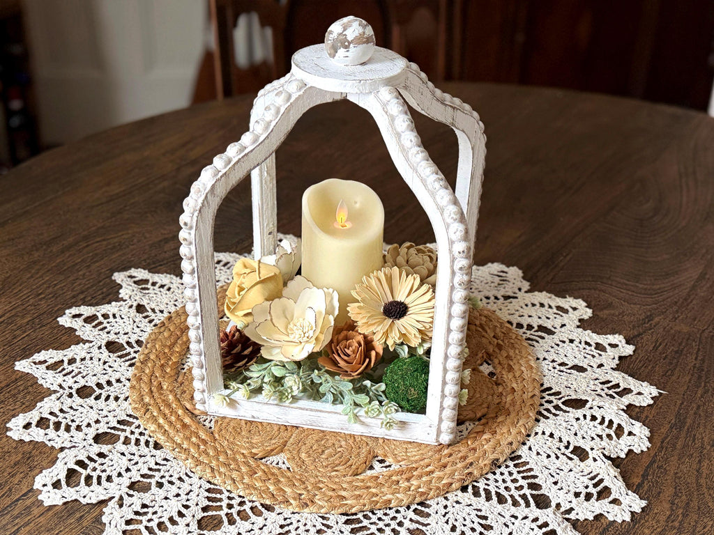 Decorative white lantern with a candle and flowers on a wooden table with a doily.